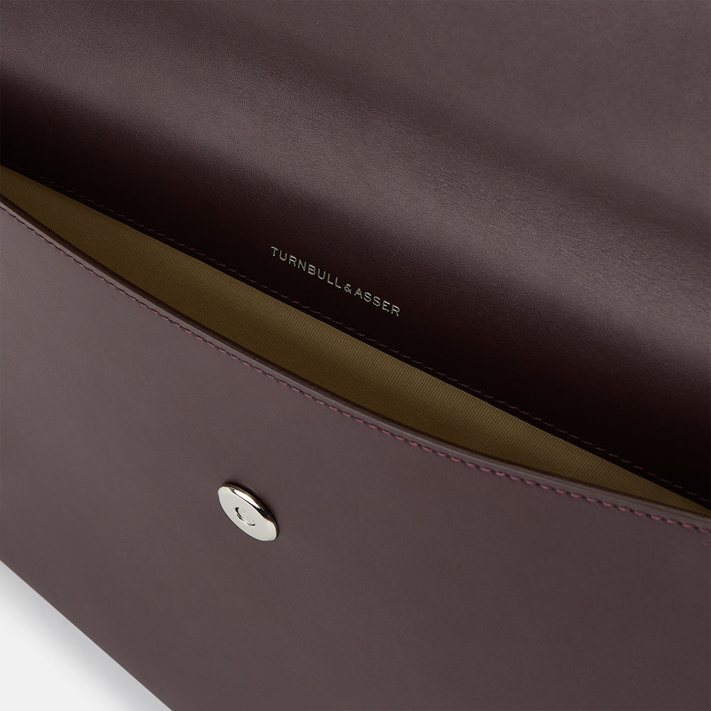 The Turnbull & Asser Burgundy Leather Snap-Fasten Pouch is a sleek, envelope-style laptop accessory with a smooth finish, curved flap, and elegant snap closure, ideal for business travel against a white background.