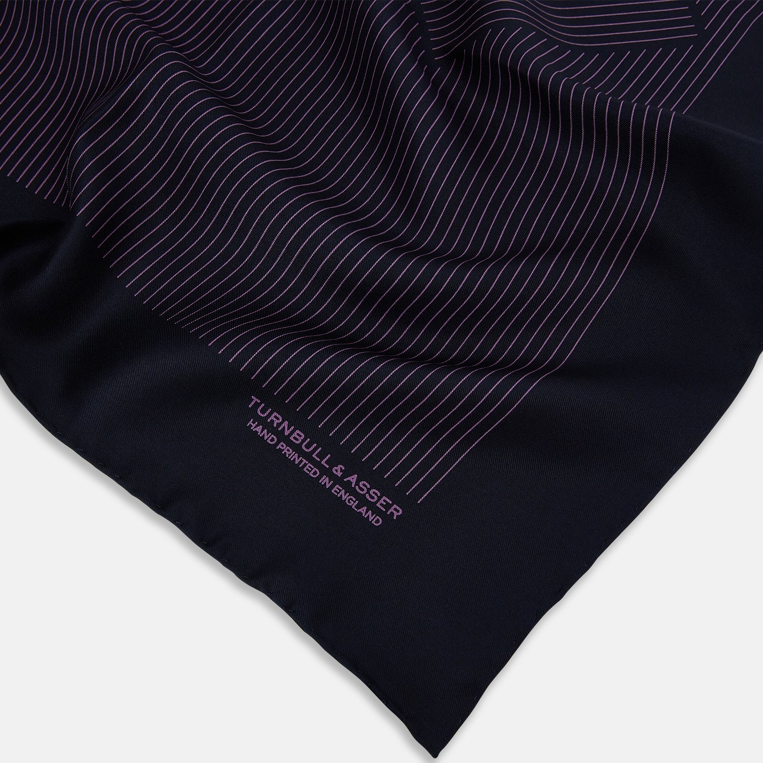 View 2 of product A navy silk pocket square with thin, even purple stripes and hand-rolled edges from Turnbull & Asser, featuring "Turnbull & Asser Hand Printed in England" printed in the corner.
