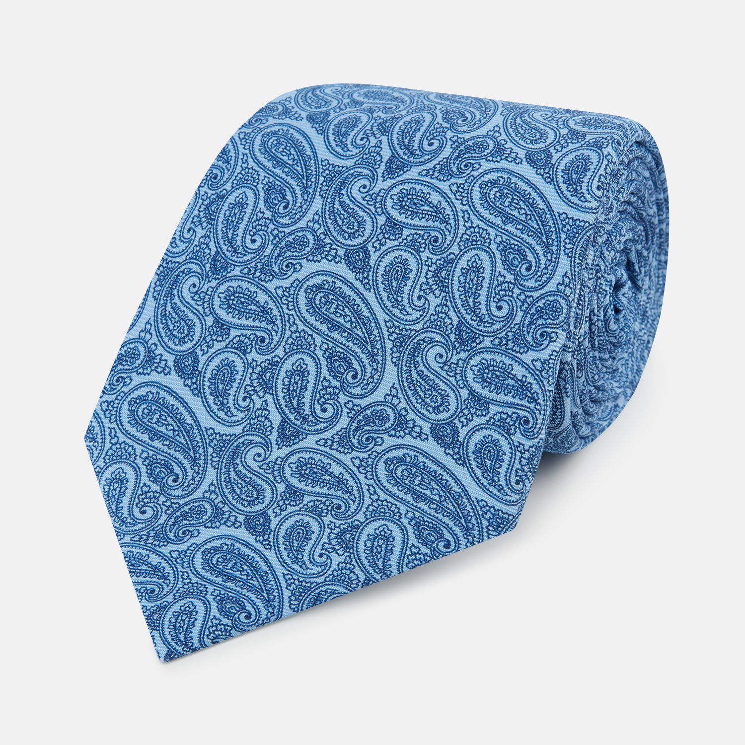 View 1 of product The Turnbull & Asser Blue Paisley Printed Silk Tie features a blue paisley pattern on a light blue background and is shown rolled up on a plain white surface.