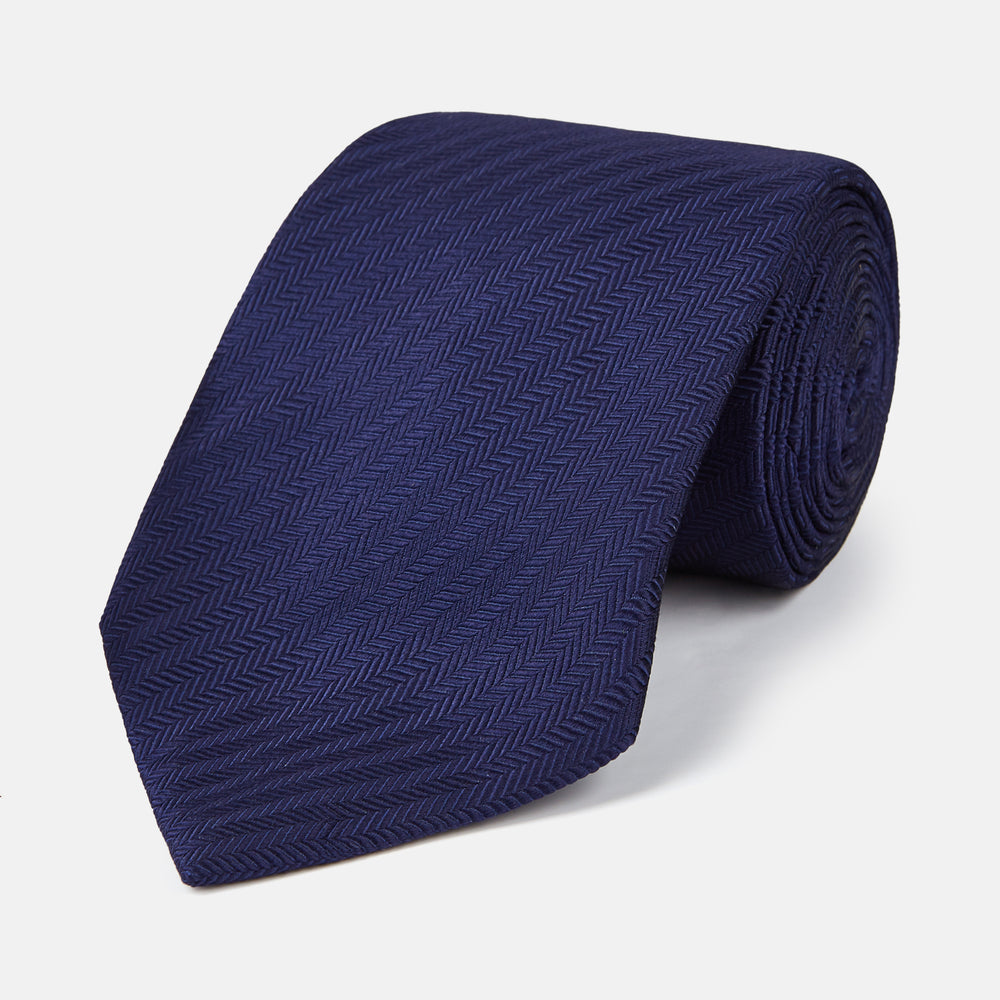The PLAIN HERRINGBONE NAVY TIE by Turnbull & Asser, crafted from silk with a subtle pattern, is elegantly rolled and displayed against a plain white background.
