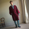 View 1 of product A person stands indoors by a window, wearing the Turnbull & Asser Burgundy Cotton-Cashmere Kimono Gown over striped pajamas, with matching slippers, next to cream curtains and a wooden chair against a pale wall.