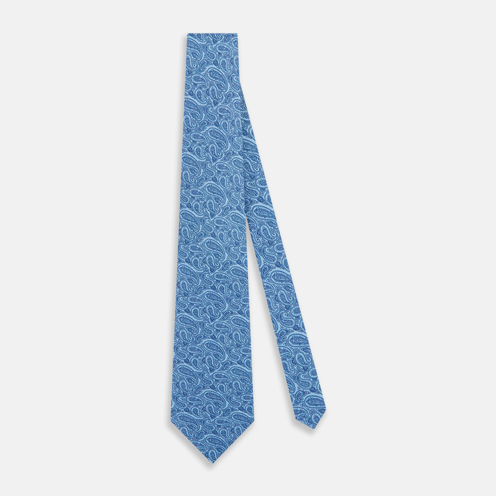 The Turnbull & Asser Blue Paisley Printed Silk Tie features a blue paisley pattern on a light blue background and is shown rolled up on a plain white surface.