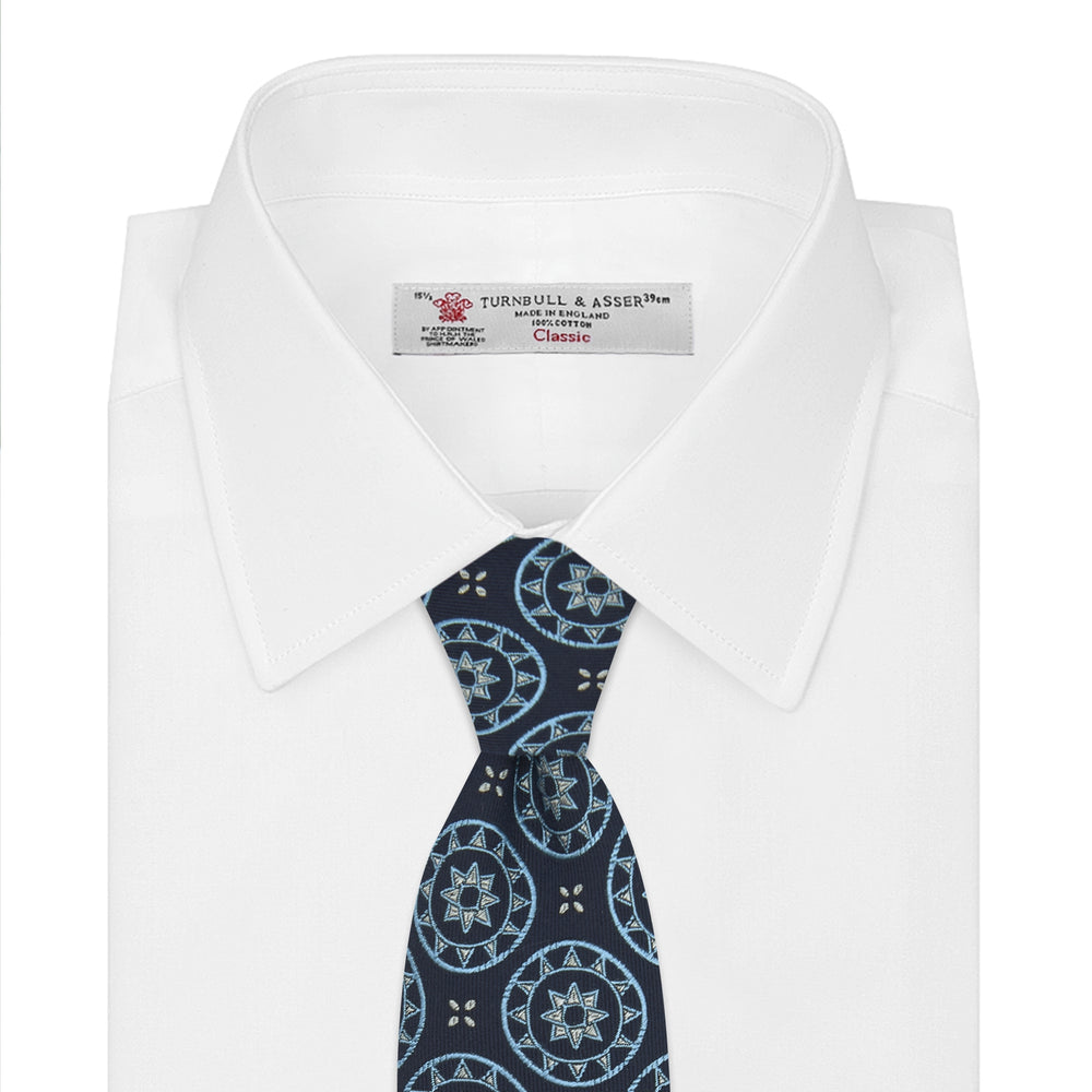 The Jacquard Silk Geometric Tie by Turnbull & Asser features a navy blue base with light blue and white star and circle motifs, shown rolled on a white background.