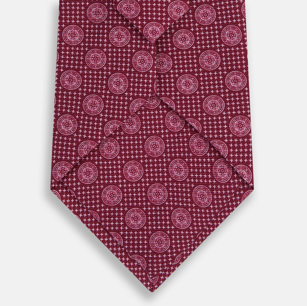 The Turnbull & Asser Burgundy Medallion Printed Silk Tie features small circular medallions and subtle light pink diagonal lines, crafted from fine English silk and displayed on a plain white background.