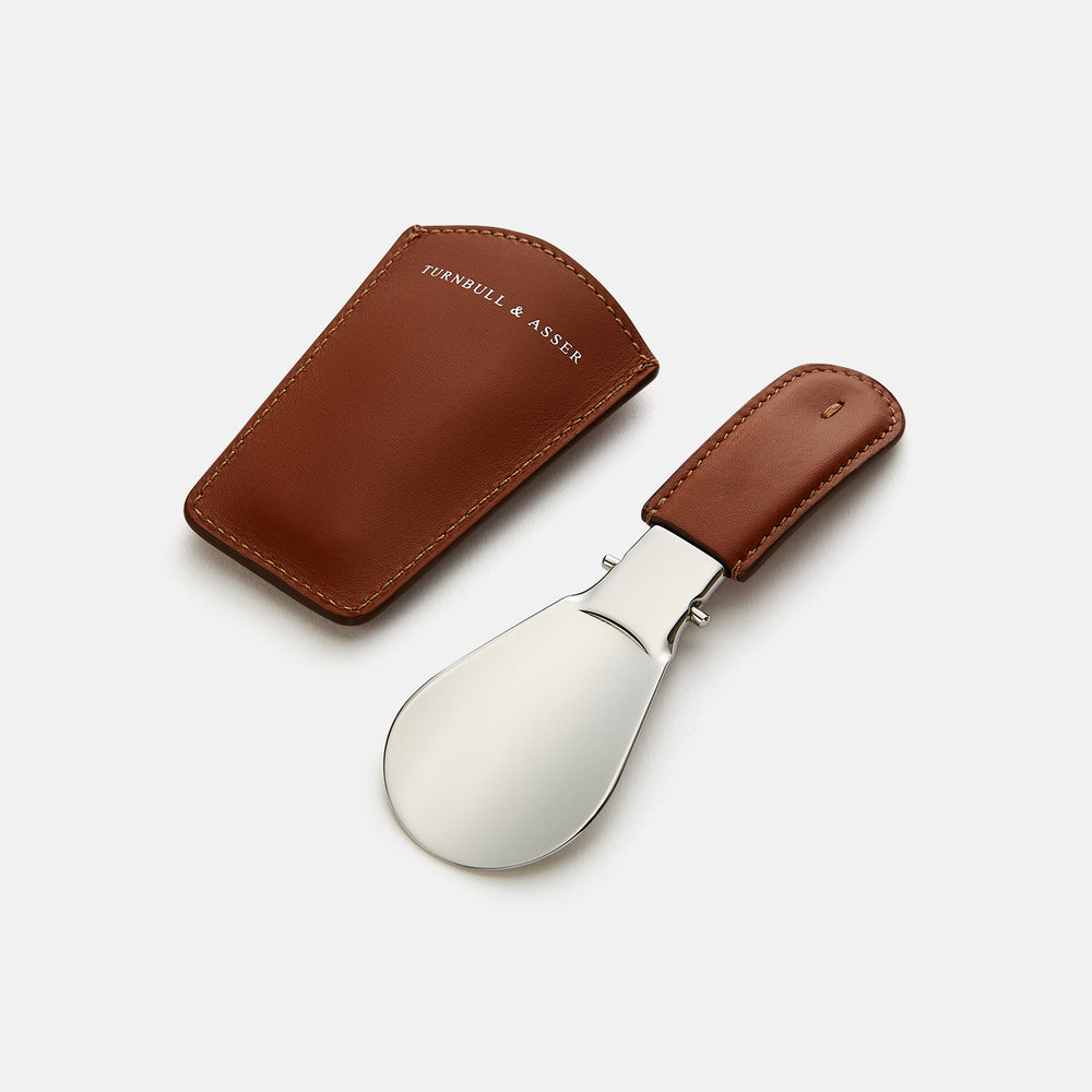 The Turnbull & Asser Camel Travel Shoe Horn features a wide, shiny silver blade and a brown Italian leather handle, displayed on a plain light background.