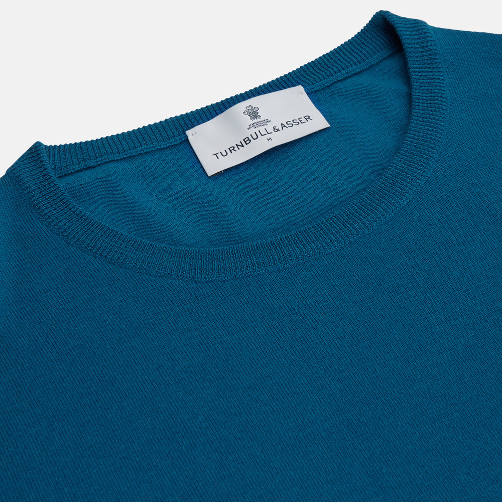 A man with light skin and short brown hair stands against a plain background, wearing the Turnbull & Asser Ocean Blue Merino Kingston Crew Neck Jumper and cream-colored pants, looking directly at the camera with a neutral expression.