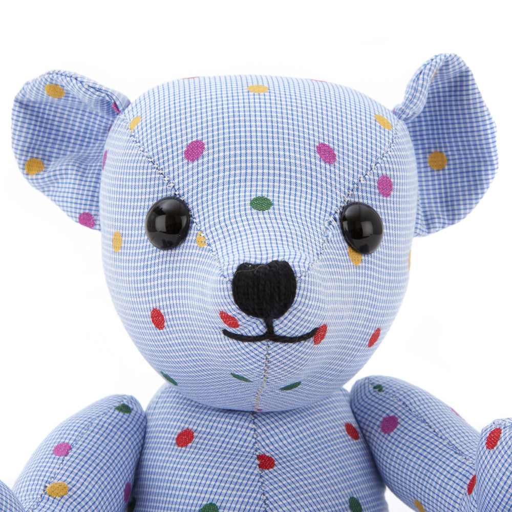 The Turnbuline Cotton Spot Teddy Bear by Turnbull & Asser is a blue gingham bear with colorful polka dots, black button eyes, a black nose, and a white leg label, shown sitting upright in profile facing left.