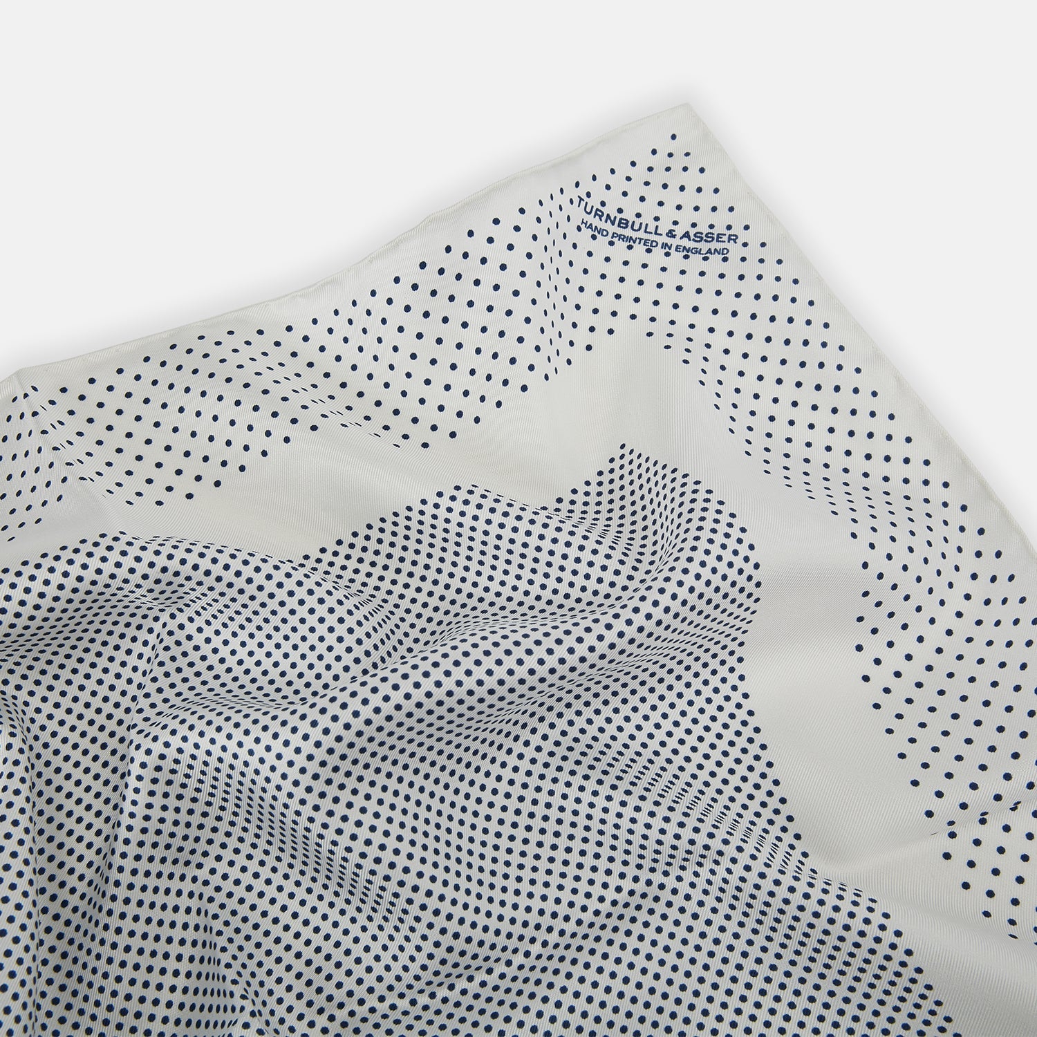 View 2 of product The Turnbull & Asser Cream Dot Silk Pocket Square features a neatly folded pure silk design with a border and center pattern of small navy blue polka dots, displayed on a plain white background.