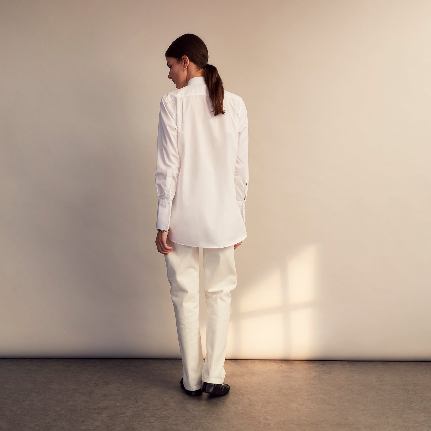 View 5 of product A person with curly hair wears the Turnbull and The Deck White Cotton Minimalist Shirt by Turnbull & Asser, paired with pants. Posed against a plain background, their minimalist silhouette is highlighted in this black and white image.
