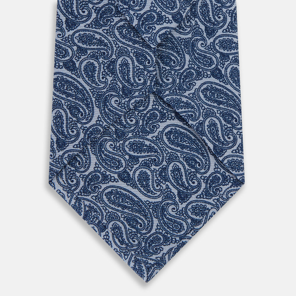 The Turnbull & Asser Pale Blue Paisley Printed Silk Tie is displayed rolled up on a white background, showcasing its intricate paisley pattern in varied blue tones.