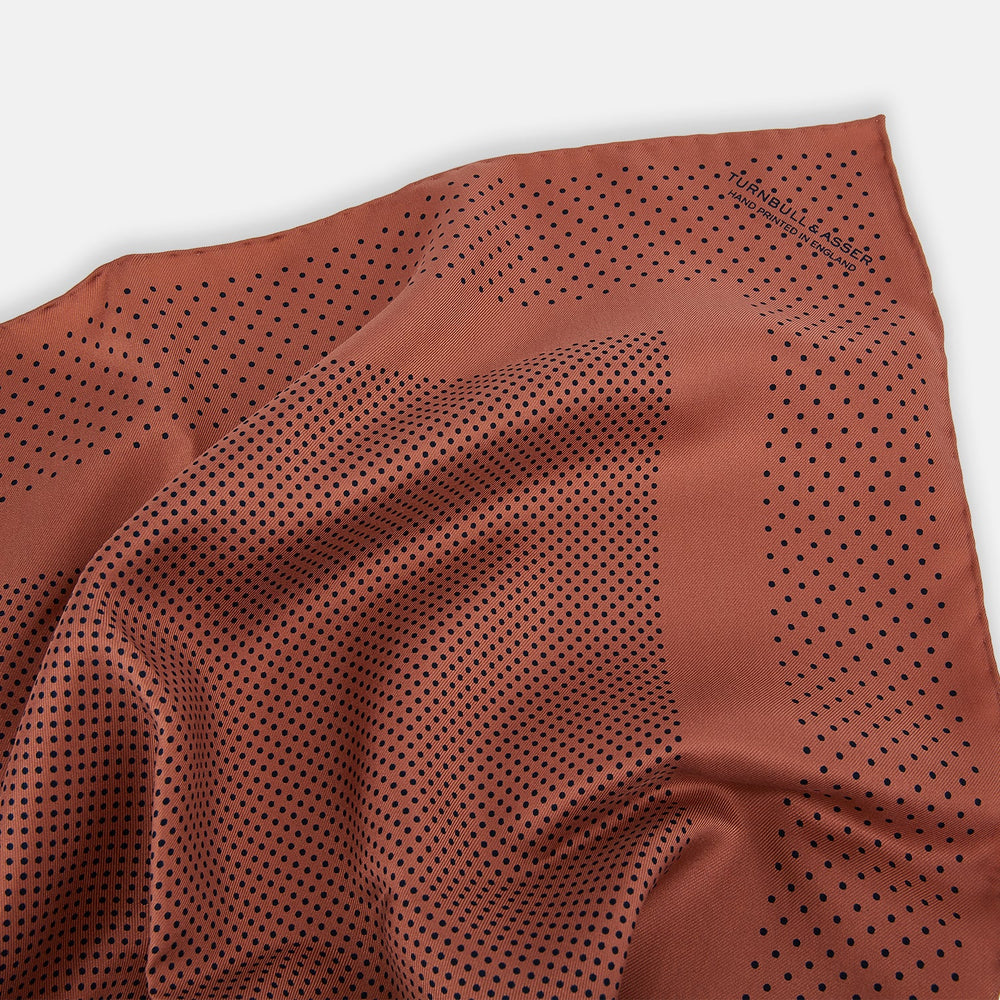 The Turnbull & Asser Blush Dot Silk Pocket Square features a rust hue with black polka dots, a bordered design, and hand-rolled edges, shown folded against a white background.