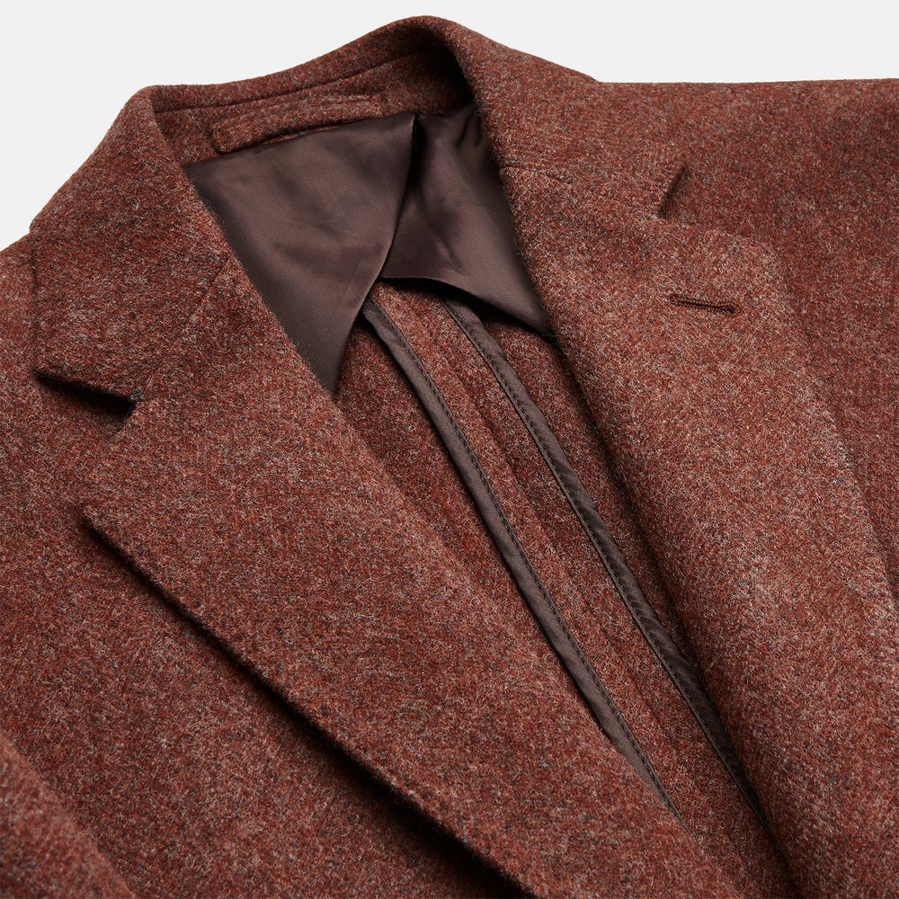 A man stands in a lush garden, hands in pockets and relaxed, wearing the Turnbull & Asser Red Lambswool Herringbone Bern Blazer, a striped dress shirt, patterned scarf, and dark trousers.
