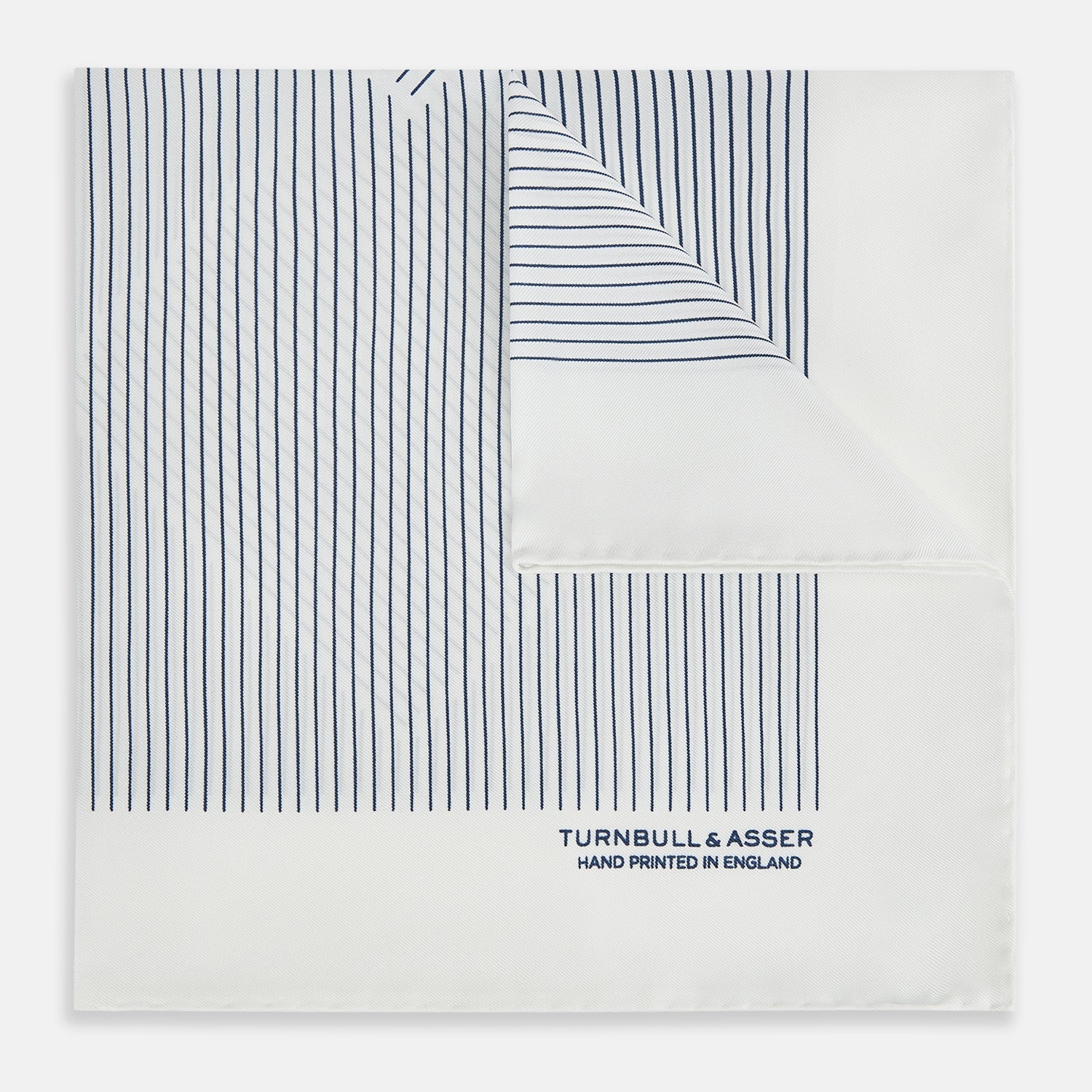 View 1 of product The White Stripe Silk Pocket Square by Turnbull & Asser features vertical navy pinstripes and “TURNBULL & ASSER HAND PRINTED IN ENGLAND” in blue at the lower right, with one corner neatly folded for a classic British look.