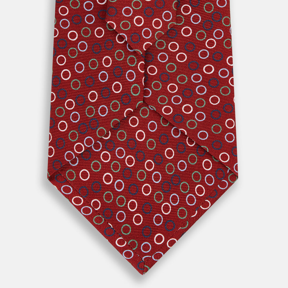 The Pink and Navy Circle Red Printed Silk Tie by Turnbull & Asser showcases English silk craftsmanship with a pattern of multicolored circles on rich red silk, elegantly displayed against a plain white background.