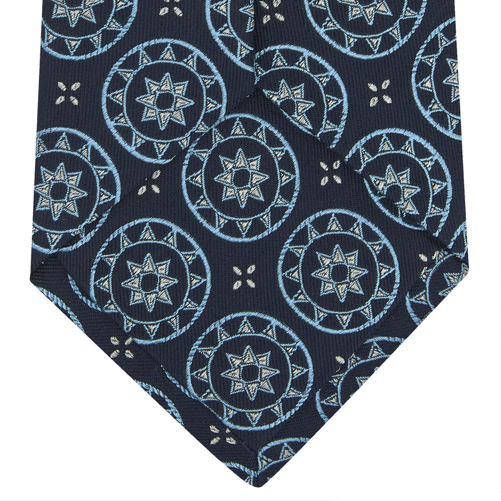 The Jacquard Silk Geometric Tie by Turnbull & Asser features a navy blue base with light blue and white star and circle motifs, shown rolled on a white background.