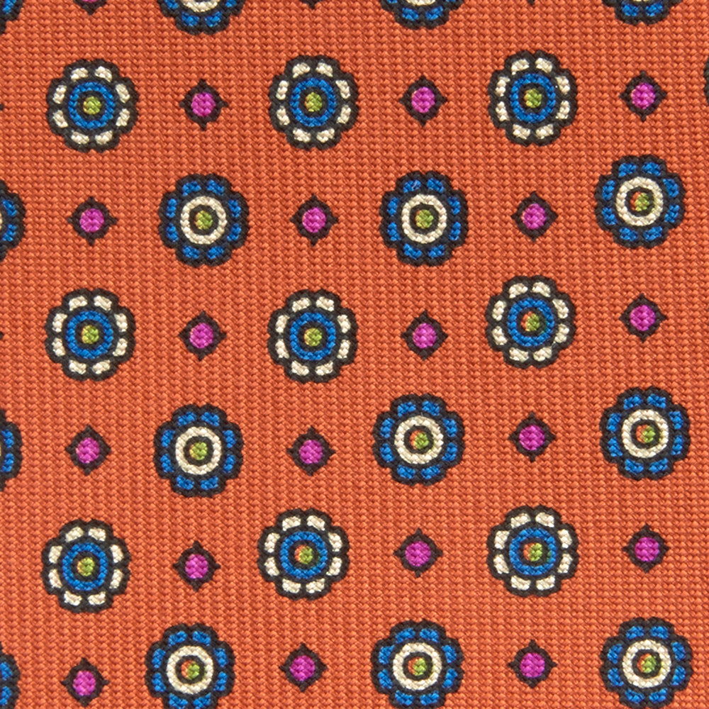 The Great Gatsby Orange Printed Silk Tie by Turnbull & Asser, from the Legends collection, features a repeating pattern of small blue, white, and yellow floral motifs—an ideal summer accessory—shown rolled on a white background.