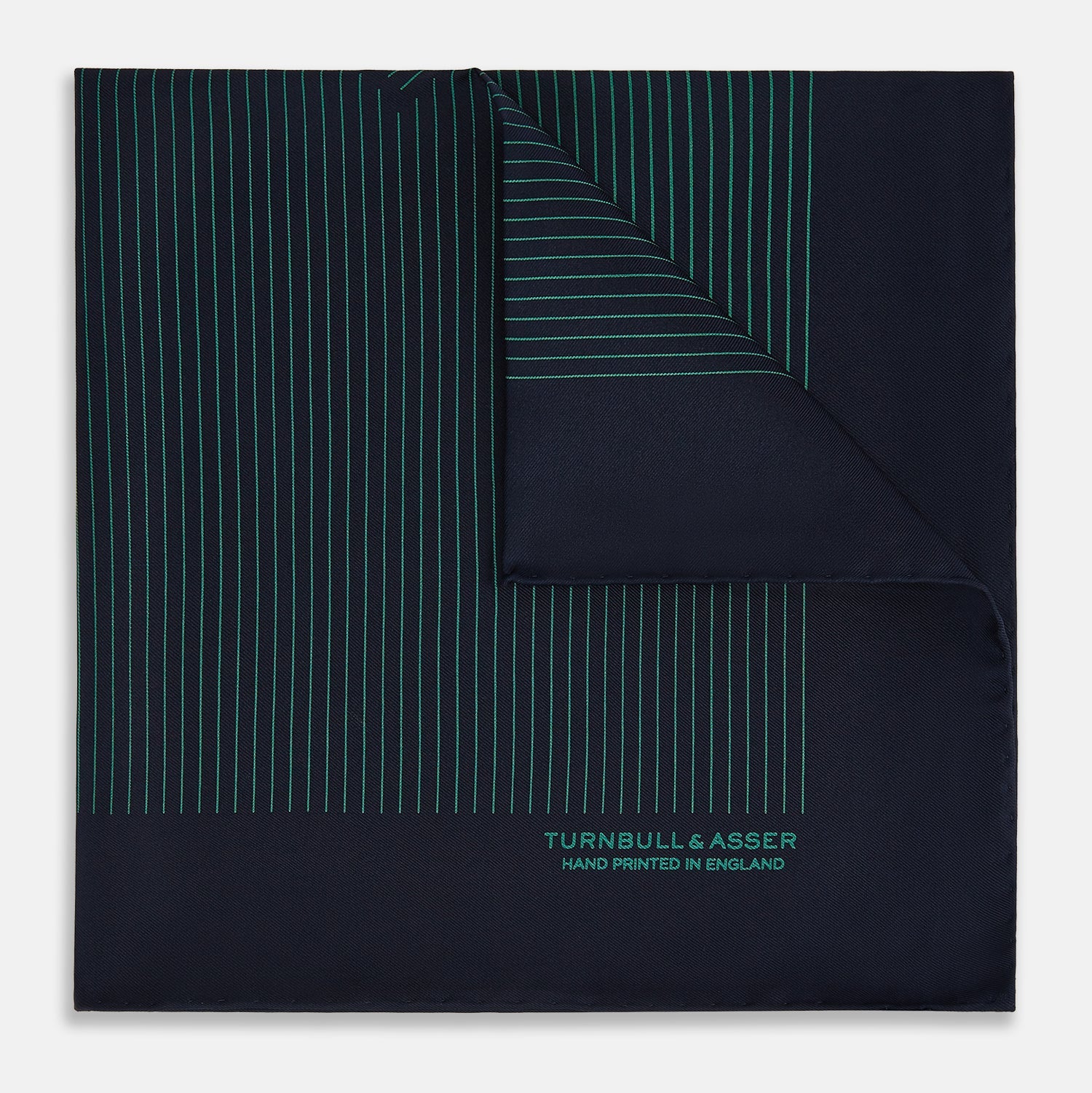 View 1 of product The Turnbull & Asser Navy and Hunter Green Stripe Silk Pocket Square features thin green stripes, hand-rolled edges, and "TURNBULL & ASSER HAND PRINTED IN ENGLAND" in green on the bottom right corner.