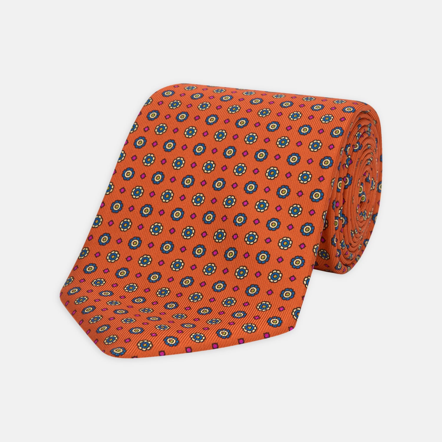 View 1 of product The Great Gatsby Orange Printed Silk Tie by Turnbull & Asser, from the Legends collection, features a repeating pattern of small blue, white, and yellow floral motifs—an ideal summer accessory—shown rolled on a white background.