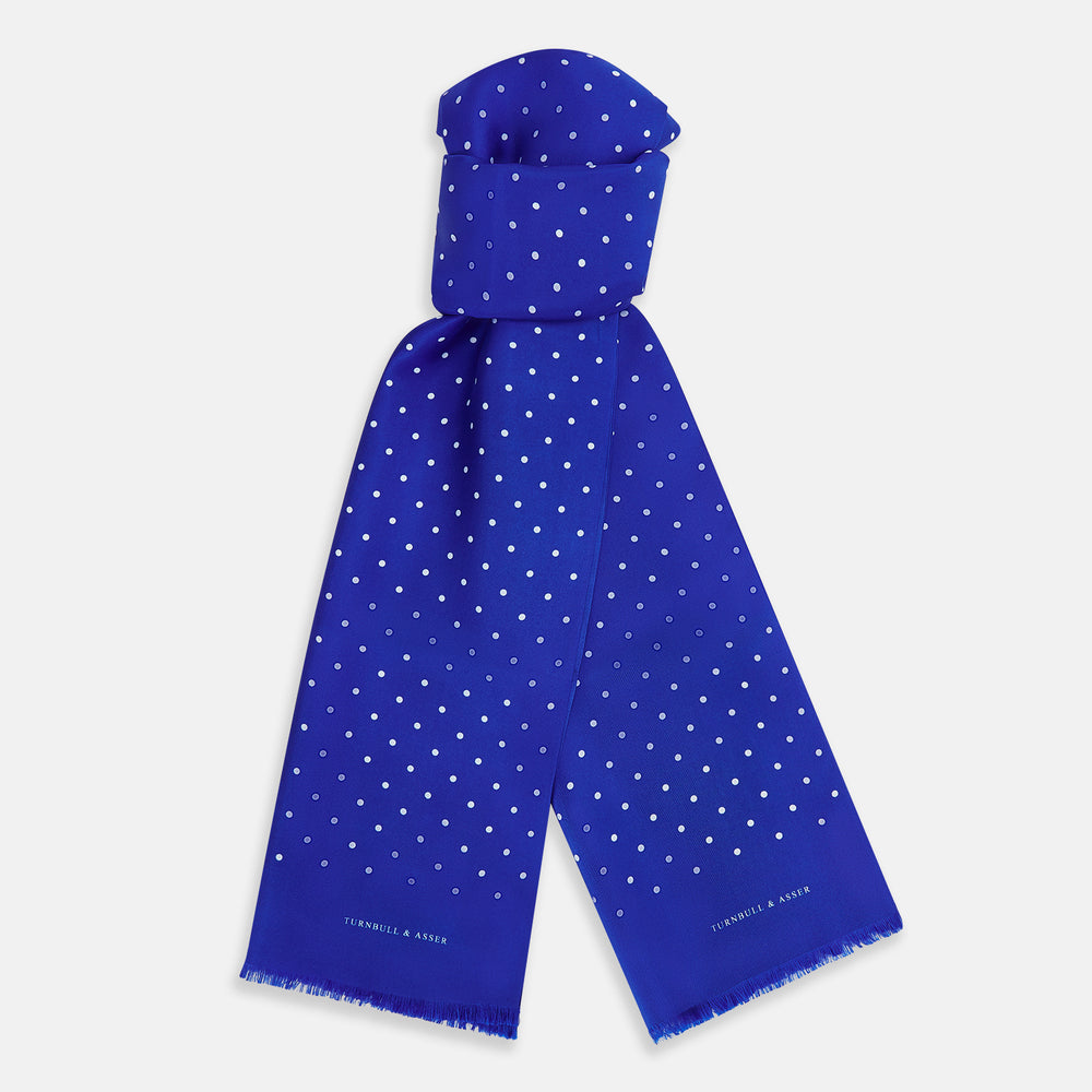 The Turnbull & Asser Royal Blue and White Spotted Silk Scarf features evenly spaced small white dots, fringed edges, and the brand’s name printed near the ends.