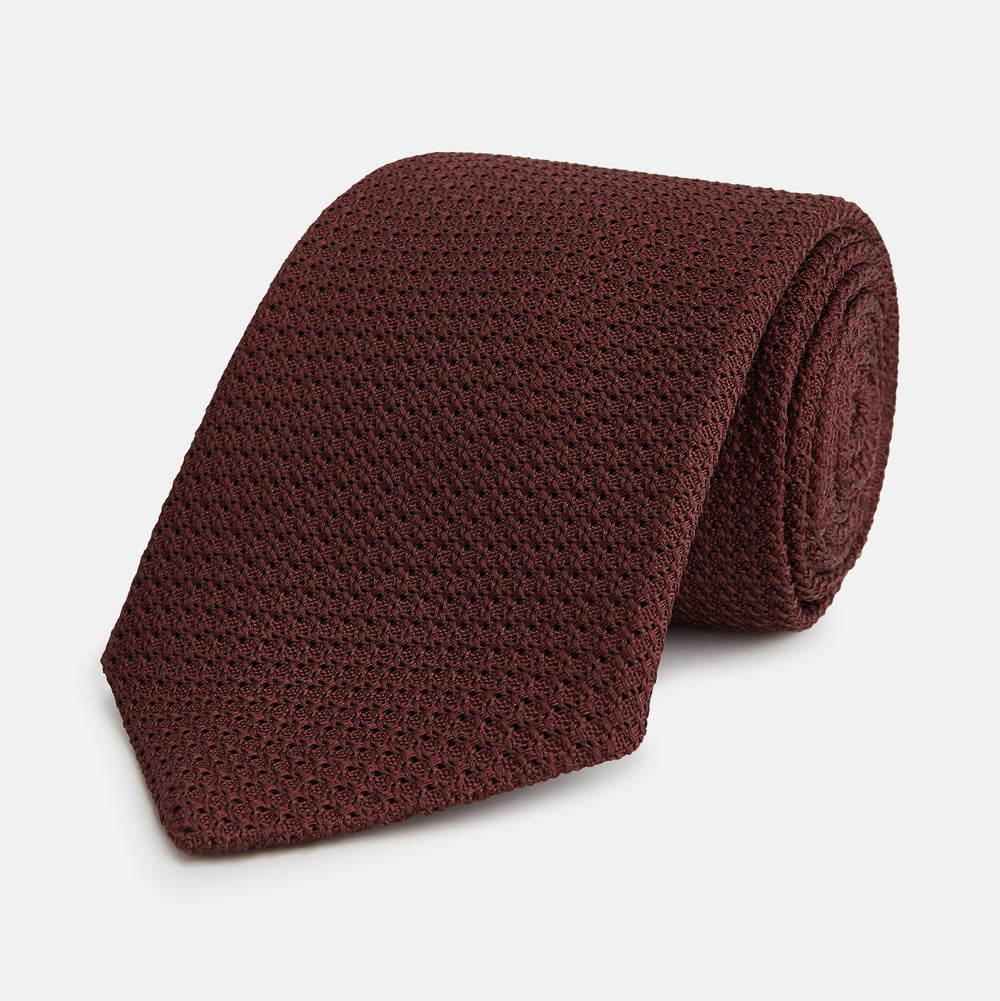 The Turnbull & Asser Ruby Red Knitted Tie, shown rolled-up against a white background, is a textured silk accessory that adds versatile sophistication to any gentleman's wardrobe.