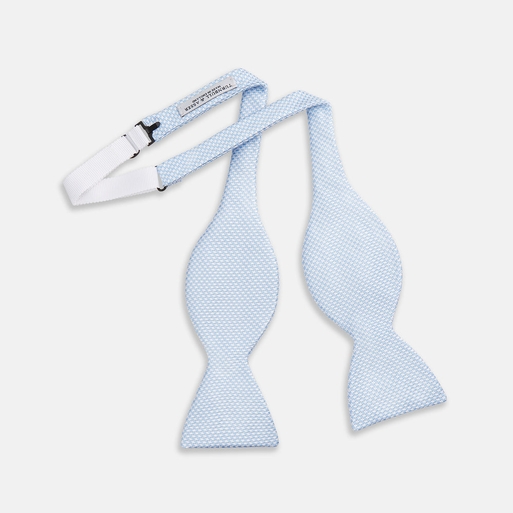 The Turnbull & Asser Pale Blue and White Houndstooth Silk Bow Tie features a refined textured pattern, making it an elegant formalwear accessory against a crisp white background.