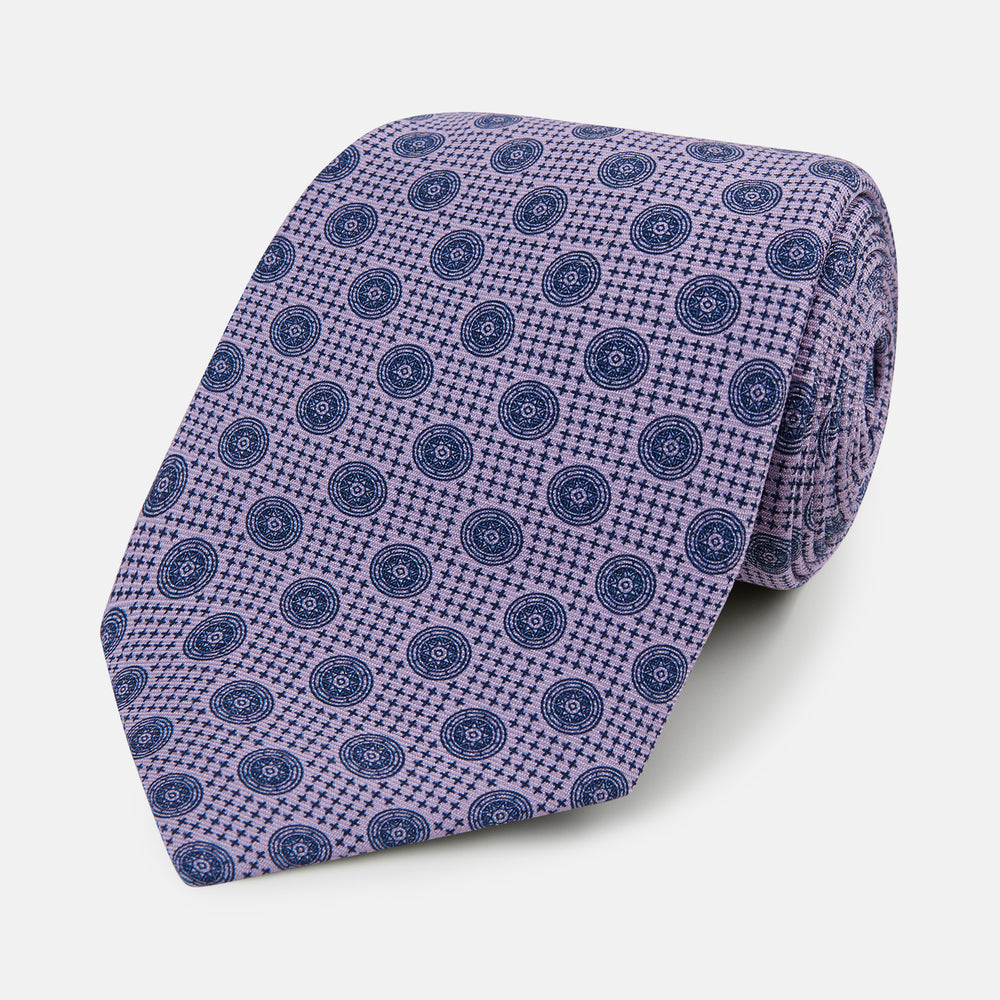 The Turnbull & Asser Lilac Medallion Printed Silk Tie is crafted from English silk with a geometric motif of small blue circles and a subtle checkered background.