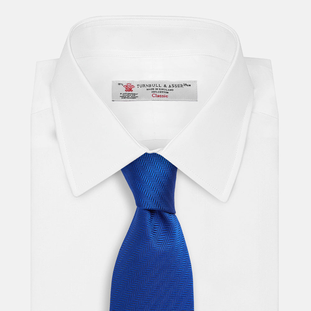 The Turnbull & Asser Plain Herringbone Cruise Tie in bright blue, shown rolled up on a white background, features a subtle chevron pattern—an essential finishing touch for your smart look.