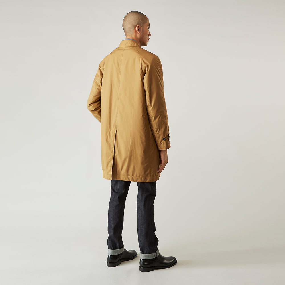 A person wears the Turnbull & Asser Camel Quintern Sealup Raincoat with Detachable Liner, paired with dark pants, standing against a plain light gray background, showing the sleek technical rainwear style with one hand in a pocket.
