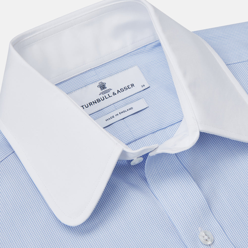 The Turnbull & Asser Micro Blue Check with White Collar and Cuff Shirt, a light blue long-sleeve style, features contrasting white collar and cuffs, buttoned down the front and laid flat on a white background.