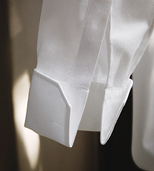 Close-up of a white dress shirt cuff with a textured fabric pattern, featuring angular, sharp edges and detailed stitching, against a softly lit background.