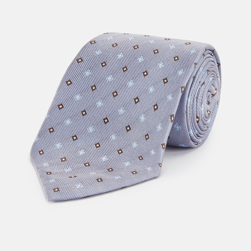 The Pale Blue Multi Floret Silk Tie by Turnbull & Asser features light purple silk with a pale blue floret pattern and small brown squares—perfect for pairing with a classic Turnbull & Asser shirt.