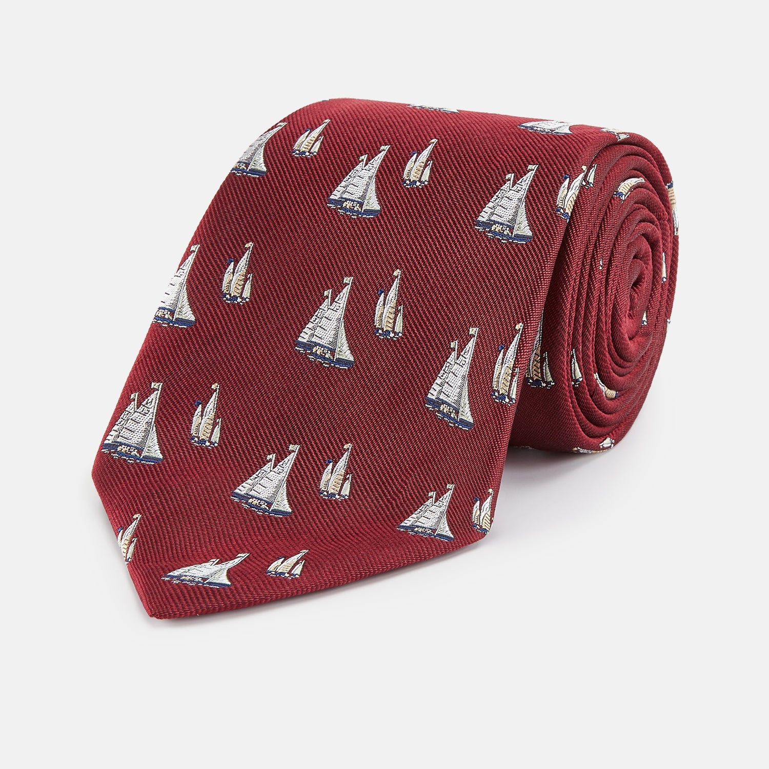 View 1 of product The Turnbull & Asser Burgundy Racing Yachts Silk Tie features a nautical sailboat pattern in white, blue, and brown on burgundy silk, shown rolled up against a plain white background.