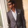 View 1 of product A man in glasses and a Turnbull & Asser Brown & Blue Check Merino Barrington Blazer, detailed with a colorful pocket square, stands thoughtfully indoors by patterned curtains, gazing out the window.