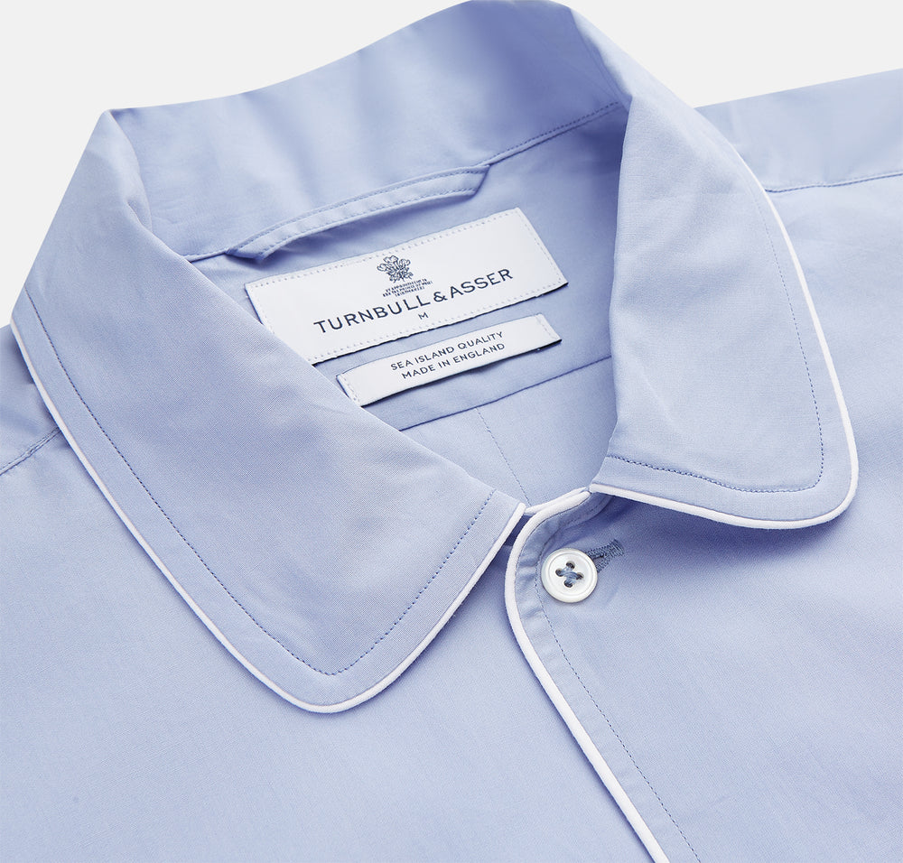 The Turnbull & Asser Blue Piped Sea Island Quality Cotton Nightshirt features light blue sea island cotton, a collared neckline with white piping, a front pocket, partial button placket, and a curved high-low hem for classic nightshirt style.