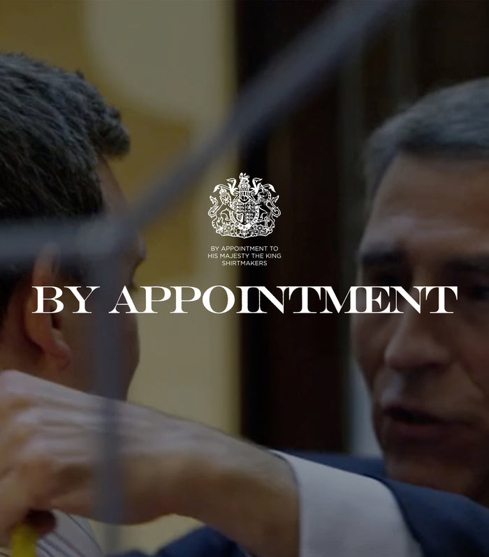 A tailor measures a mans shoulder with a yellow measuring tape. Overlaid text reads BY APPOINTMENT with a royal crest above, indicating a royal warrant for shirtmakers.
