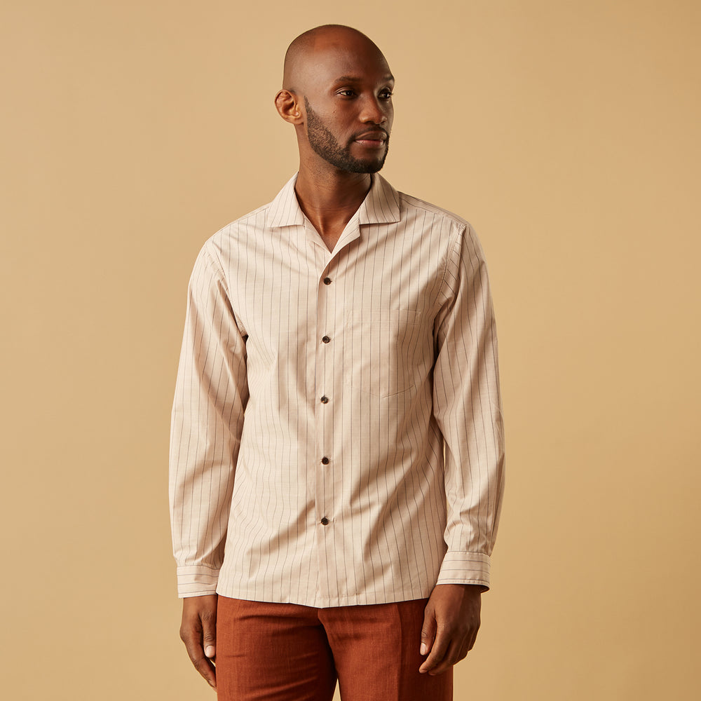 An older man in a light hat, Turnbull & Asser Beige Stripe Cotton Holiday Fit Shirt with Revere Collar & Single Button Cuff, and white pants leans casually against a tall concrete pillar outdoors, greenery blurred behind him.