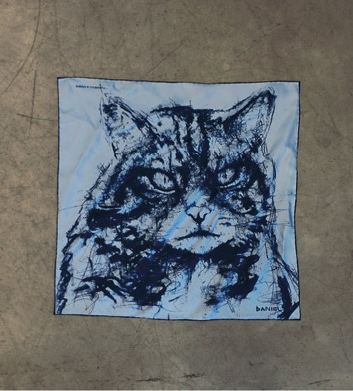 A square cloth with a blue, sketch-style drawing of a cats face, featuring intense eyes and bold lines, is laid flat on a plain, gray surface.