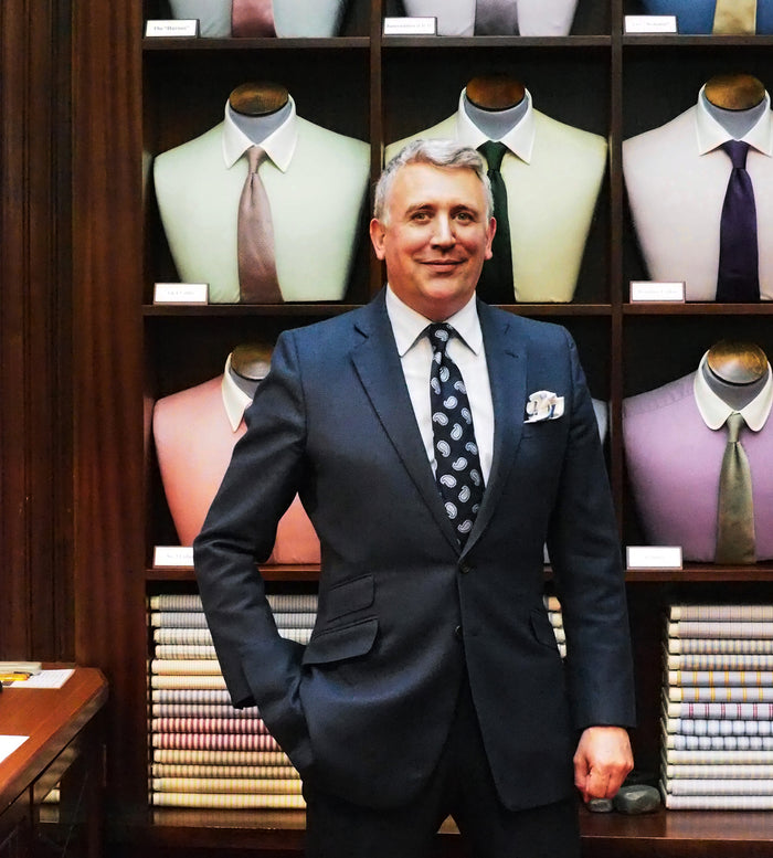 A man in a dark suit stands smiling in front of shelves displaying mannequin torsos dressed in pastel shirts and ties, with folded fabrics stacked below.