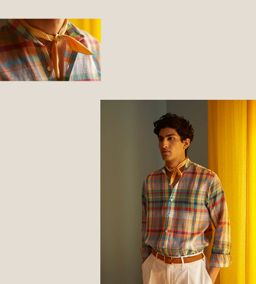 A man wearing a colorful plaid button-up shirt, white pants, and a yellow scarf around his neck stands indoors next to a yellow curtain. A close-up of the shirt and scarf is shown in the top left corner.