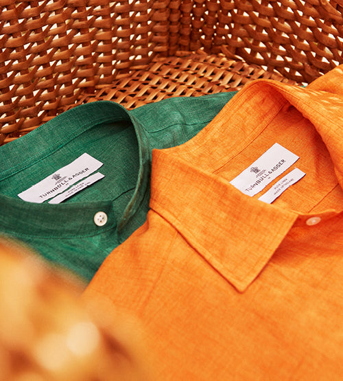 Two neatly folded shirts, one green and one orange, are displayed side by side in a woven wicker basket. Both shirts have visible labels on the inside of the collars.