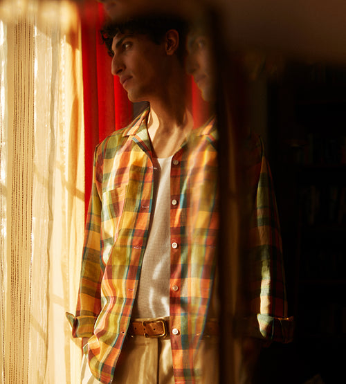 A person in a colorful plaid shirt stands by a sunlit window, looking outside with a thoughtful expression. Warm sunlight and sheer curtains create a soft, reflective atmosphere, with part of the reflection visible in a mirror.