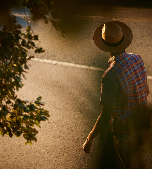 A person wearing a straw hat and plaid shirt walks along a sunlit road, partially shaded by tree branches. The scene is viewed from above and behind.