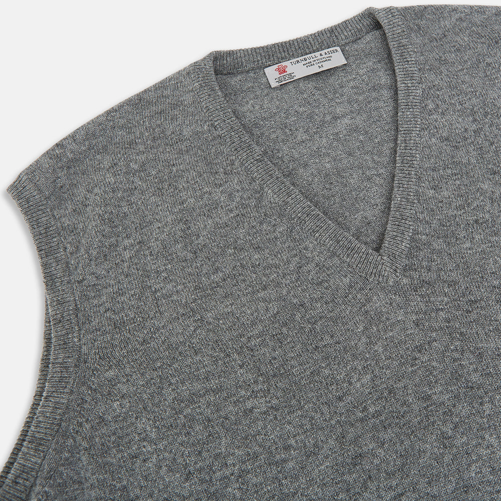 Turnbull & Asser UK’s marl grey sleeveless V-neck vest in cashmere, featuring a ribbed hem and crafted from soft knit fabric, is showcased against a plain white background.