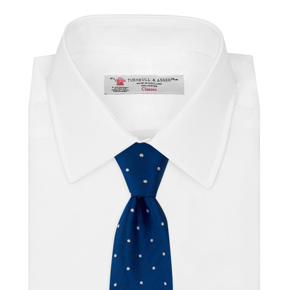 The Turnbull & Asser Royal Blue and White Small Spot Herringbone Silk Tie is displayed rolled at one end on a plain white background, showcasing its classic English silk design with subtle white polka dots.