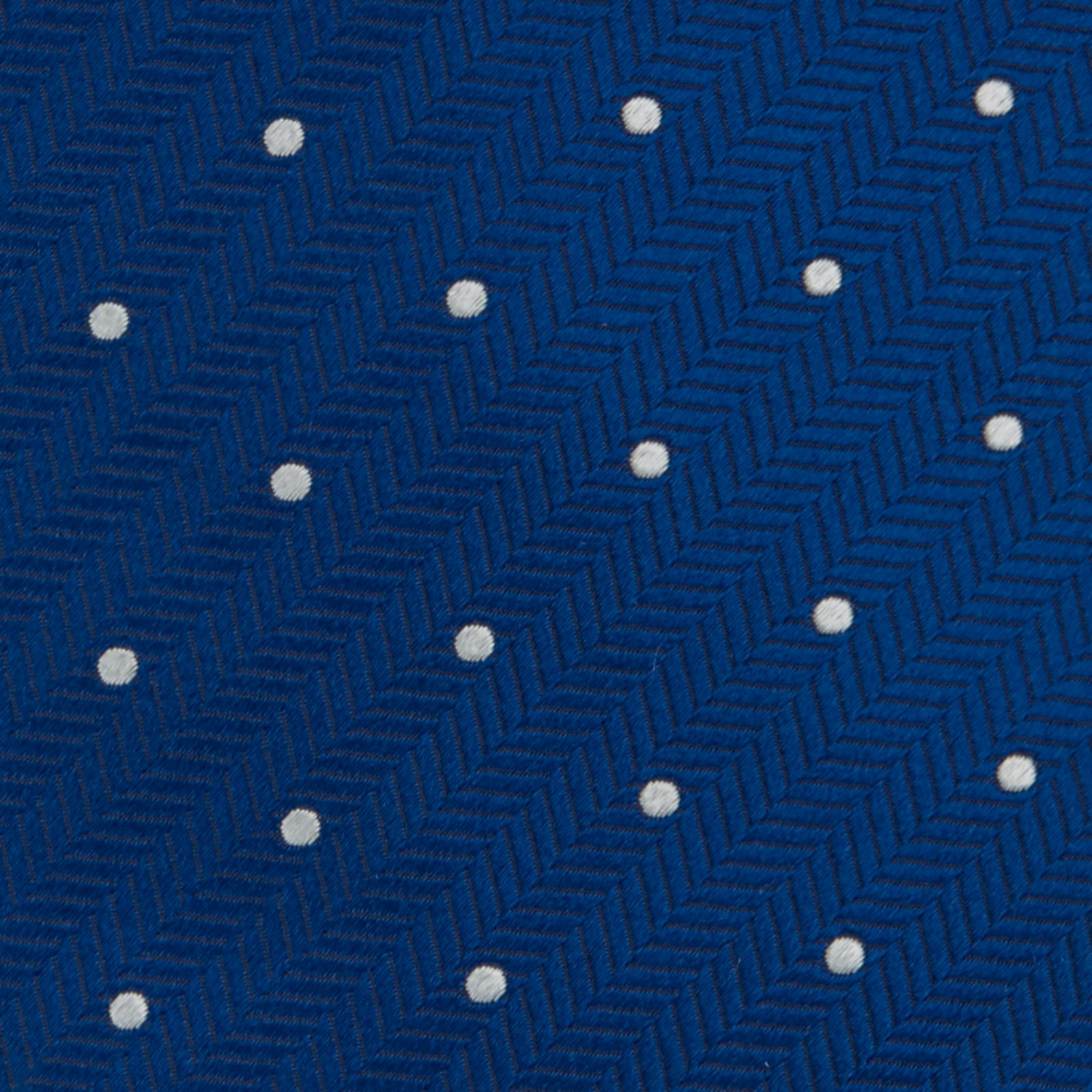 View 3 of product The Turnbull & Asser Royal Blue and White Small Spot Herringbone Silk Tie is displayed rolled at one end on a plain white background, showcasing its classic English silk design with subtle white polka dots.