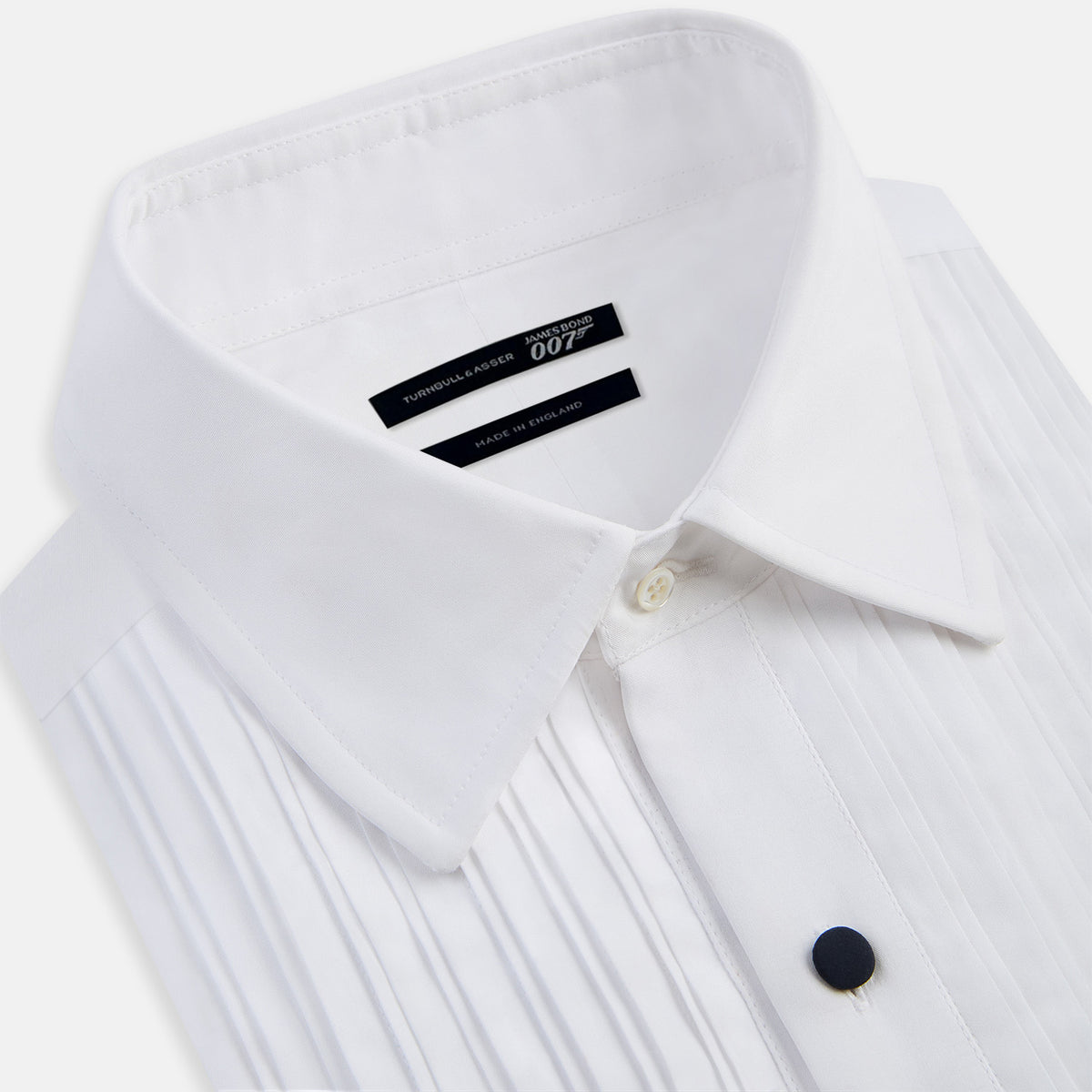 View 2 of product The Casino Royale White Dress Shirt in Cotton by Turnbull & Asser UK is a long-sleeve white dress shirt, crafted from crisp white cotton poplin with a pleated bib front, black buttons, and a classic collar, shown laid flat on a white background.