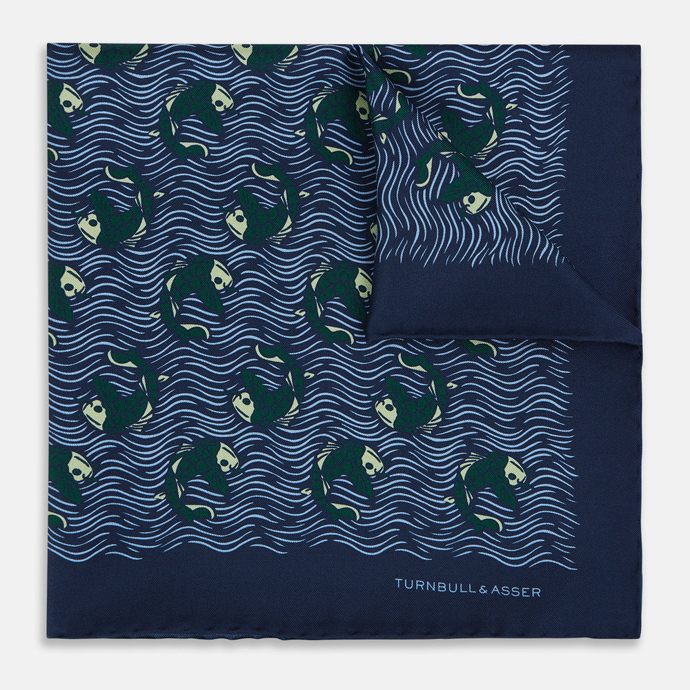 The Navy Blue Wave Silk Pocket Square by Turnbull & Asser UK showcases a playful motif of green and white fish swimming among wavy blue lines, with the brand’s name printed in white in the corner.