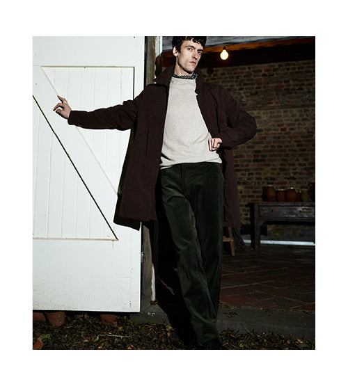 A man poses indoors wearing a dark brown coat, light beige sweater, and dark green pants. He stands with one hand resting on a white wooden door and the other in his pocket. The background features brick walls and a rustic setting.