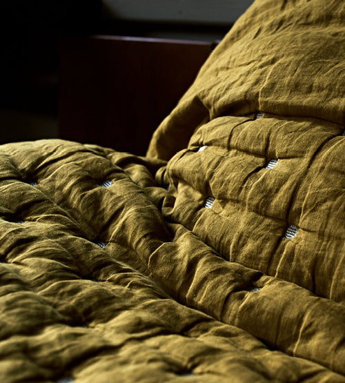 Close-up of a mustard yellow, textured, quilted blanket and pillow on a bed, with soft natural light highlighting the fabric’s wrinkled appearance and tufted details.