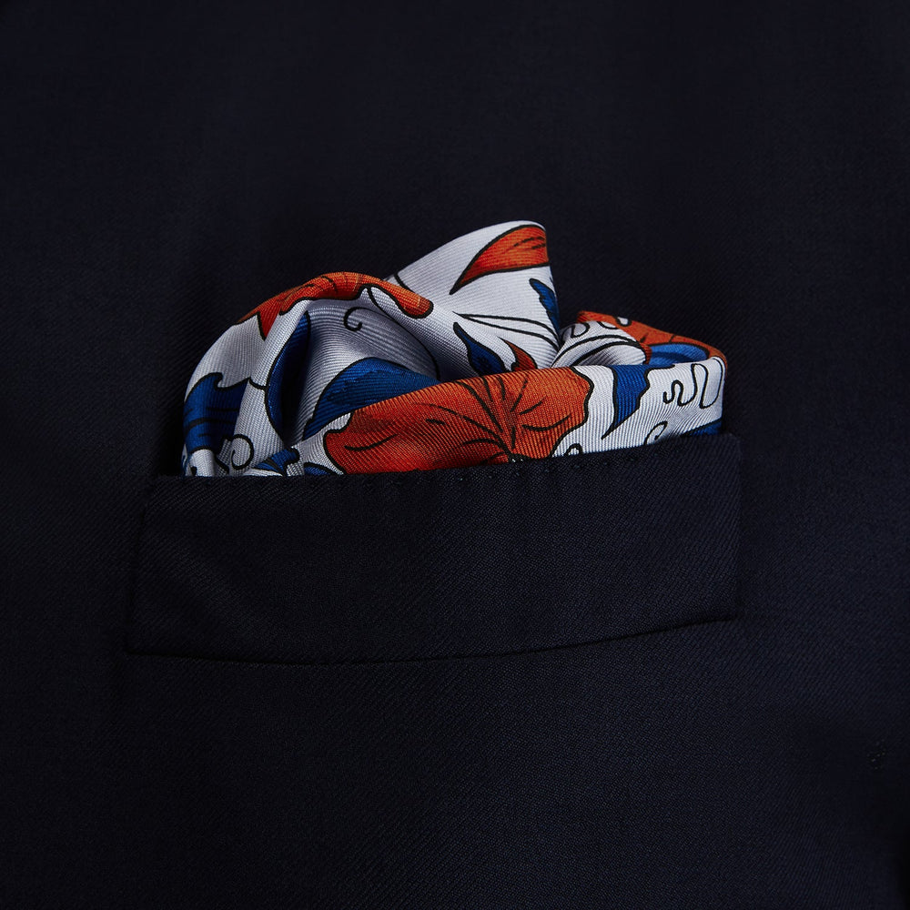 The Blue and Red Foliage Pattern Silk Pocket Square by Turnbull & Asser features intricate symmetrical leaves and flowers in red, blue, and navy on a light grey silk background, creating an elegant circular motif.